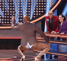 a man in a suit and high heels is squatting on a stage with a woman in a purple jacket behind him