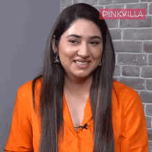 a woman wearing an orange shirt and earrings smiles in front of a brick wall with pinkvilla written on it