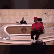 a man in a red shirt is sitting in a wheelchair on a television show .