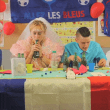 a man and a woman sit at a table with bottles of beer in front of a banner that says allez les bleus