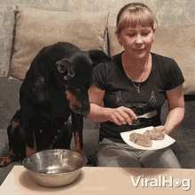 a woman sits at a table with a plate of food and a bowl of water and a dog looks on