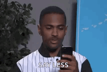 a man in a striped shirt is holding a cell phone in his hand and saying `` say less '' .