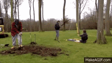 a man is digging a hole in the grass while another man sits on the ground holding a stick .