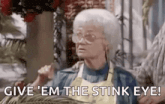 an elderly woman wearing glasses and an apron is sitting on a couch and saying `` give em the stink eye ! ''