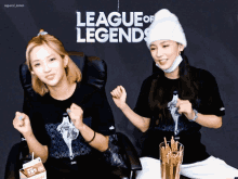 two women sitting in front of a league of legends sign