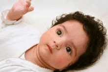 a baby with curly hair is laying on a bed and looking up at the camera .