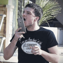 a man wearing a black shirt that says clermont lounge is holding a cell phone in his mouth