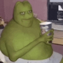 shrek is sitting at a table holding a cup of coffee and a cup of popcorn .