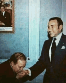 a man in a suit is kissing another man 's hand in front of a painting .