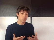 a young man in a black shirt is talking to someone while standing in front of a white wall .