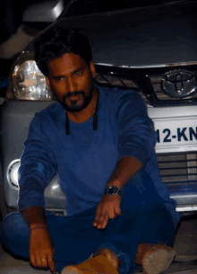 a man sits on the ground in front of a car with a license plate that says 12-kn