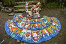 an elderly woman is sitting on the ground painting a colorful painting on a piece of cloth .