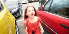 a little girl in a red dress is standing in front of a red car in a parking lot .