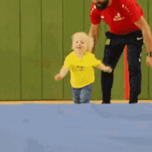 a little girl in a yellow shirt is jumping on a blue mat with a man in a red shirt standing behind her