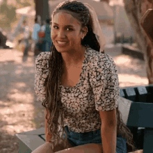 a woman with braids is sitting on a bench and smiling . she is wearing a floral shirt and shorts .