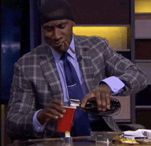 a man in a suit and tie is smoking a cigar and pouring a drink into a red cup