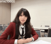a woman in a red jacket and tie sits at a table in front of a wall with the number 9791/10000 on it