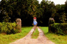 a man in a blue shirt is walking down a dirt road with a sign on the right that says ' a '