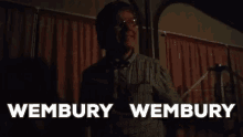 a man is standing in front of a microphone in a dark room with the words wembury wembury behind him .