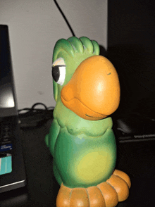 a green and orange bird figurine sits on a desk next to a laptop