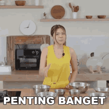 a woman in a yellow tank top is standing in a kitchen with bowls on the counter and the words penting banget on the bottom .