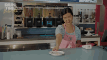 a woman in a pink apron reaches for a plate of food in a kitchen with the words run the burbs behind her