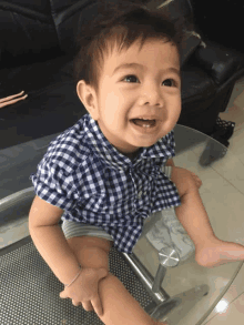 a baby wearing a plaid shirt is sitting on a glass table and smiling