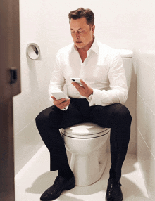 a man in a white shirt sits on a toilet looking at his phone