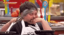 a man in an apron is holding a tong in his mouth while sitting in a kitchen .