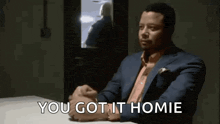 a man in a suit is sitting at a table with his hands folded and the words `` you got it homie '' .