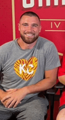 a man wearing a kc shirt is sitting in front of a red wall .