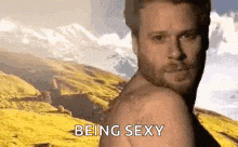 a shirtless man is standing in front of a mountain with the words `` being sexy '' written on the bottom .