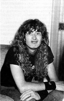 a man with long curly hair is sitting on a couch with his hands folded .