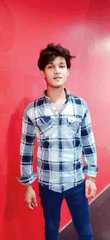 a young man standing in front of a red wall wearing a plaid shirt