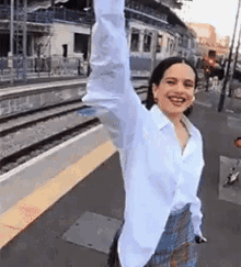 a woman in a white shirt and plaid skirt is standing on a train platform with her arms in the air .