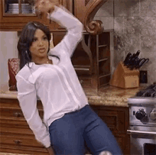 a woman in a white shirt and blue jeans is standing in a kitchen with her arm in the air .