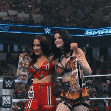 two women in a wrestling ring holding their championship belts with a sma logo in the background