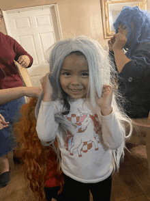 a little girl wearing a unicorn shirt is holding a wig