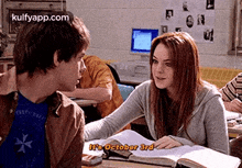 a boy and a girl are sitting at a desk in a classroom and the girl is talking to the boy .