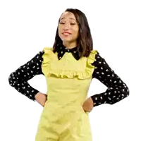a woman in a yellow dress and black polka dot shirt is standing with her hands on her hips and smiling .