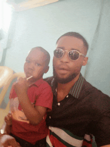 a man wearing sunglasses is holding a child who is eating a lollipop