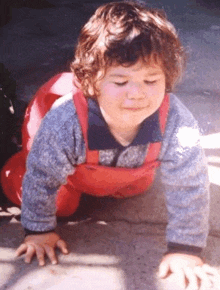 a young child wearing red overalls and a blue sweater is crawling on the ground