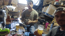 two men are drinking beer in a messy room with a box that says ventilador pie pared on it