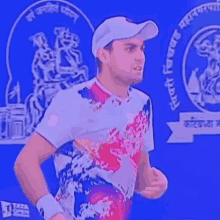 a man wearing a white hat and a colorful shirt is standing in front of a blue wall with a logo in the background .