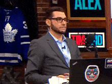 a man in a suit is sitting in front of a laptop with a toronto maple leafs jersey behind him