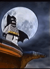 a lego batman standing on top of a building with a full moon behind him