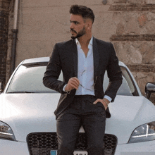 a man in a suit leans against a white car with a license plate that says ada