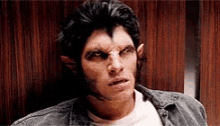 a man with a werewolf make up on his face is sitting in front of a wooden wall .