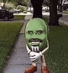 a green m & ms mascot is standing on a sidewalk holding a m & m .