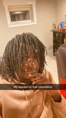 a young man with dreadlocks is sitting in a living room with his hand on his chin and a caption that says my reaction to that information